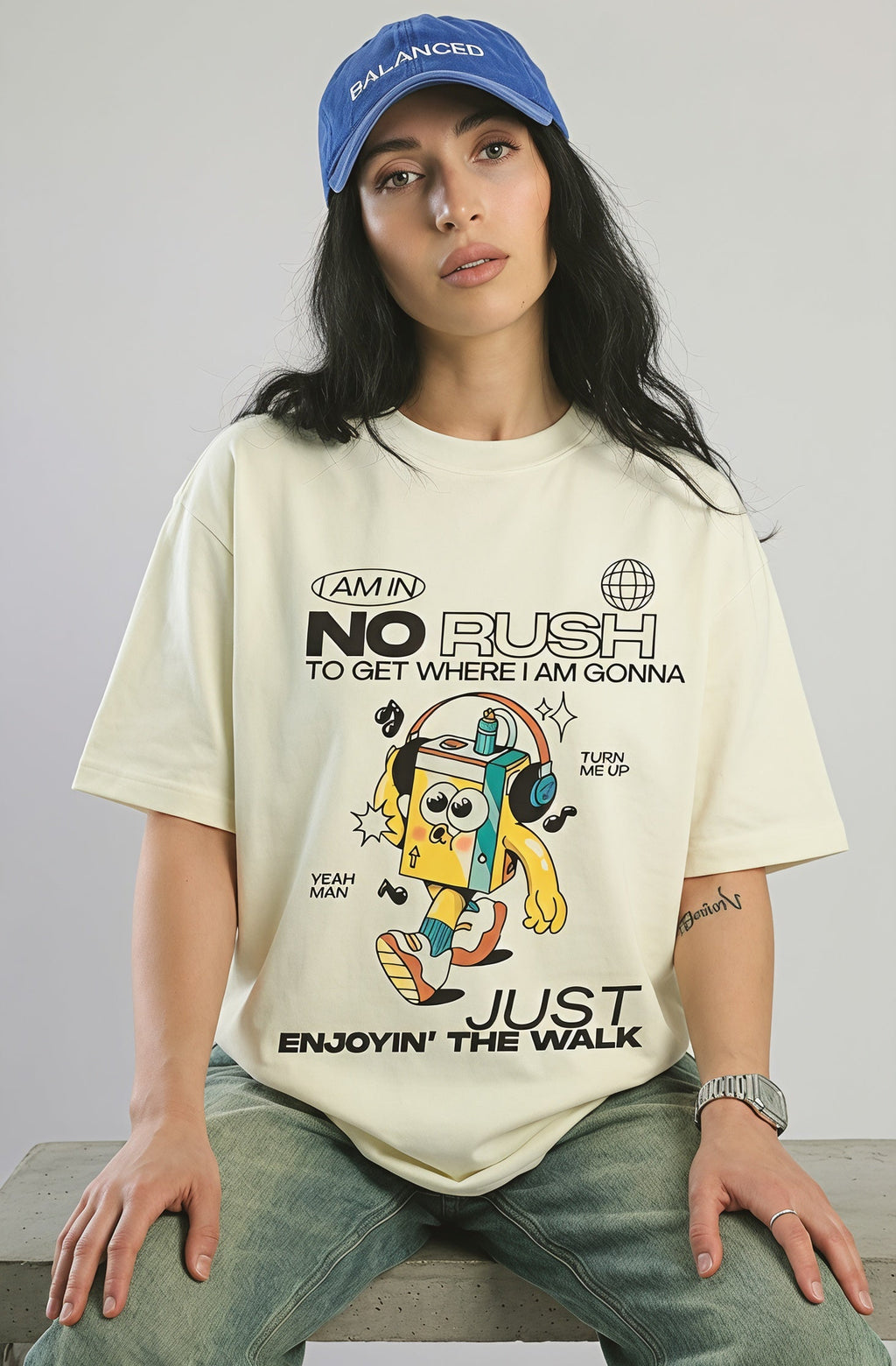 Person wearing a graphic t-shirt and blue cap sitting on a white box against a plain background