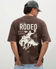 Man wearing a brown t-shirt with a rodeo graphic and text on the back, standing against a plain background.