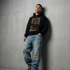Man wearing a black hoodie with graphic design and ripped blue jeans against a gray wall.